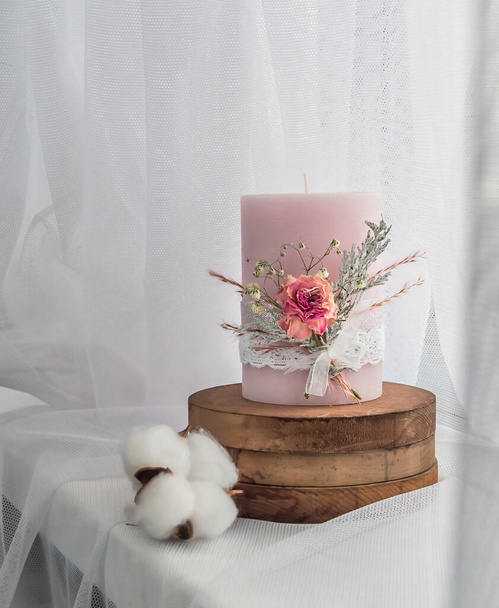 Deze handgemaakte roze kaars is versierd met gedroogde bloemen - roos, varens en wit kant op houten ronde benodigdheden. Tegen de achtergrond van witte tule door het raam - Foto, afbeelding