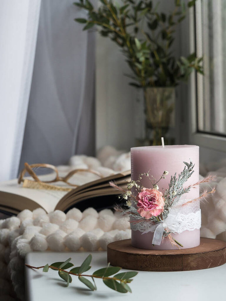 Glasses in an open book on a cozy windowsill with candles, a blanket, a wooden stand, a vase of water and green twigs by the window. Creating a cozy atmosphere in the home. - Photo, Image