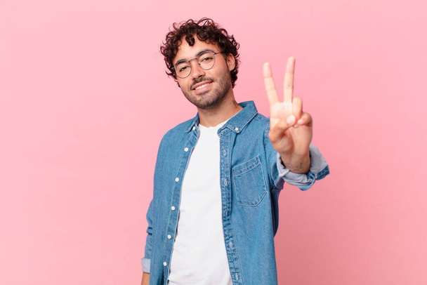 hispanic handsome man smiling and looking happy, carefree and positive, gesturing victory or peace with one hand - Foto, Imagen