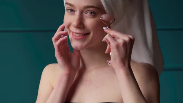 Piękna piegowata kobieta masuje twarz wałkiem derma uśmiechającym się do kamery po kąpieli - Materiał filmowy, wideo