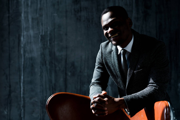 An investor from Nigeria stands in a formal suit smiling and leaning his elbows on a chair with his hands - Foto, Imagen