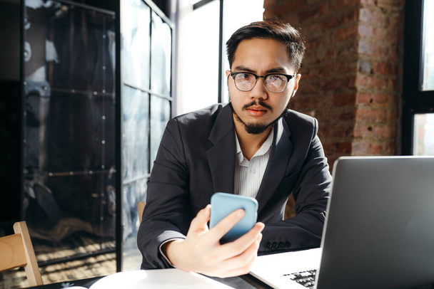 Vážný asijský podnikatel sedí u stolu s notebookem a dívá se na telefon, zatímco ho drží v ruce - Fotografie, Obrázek