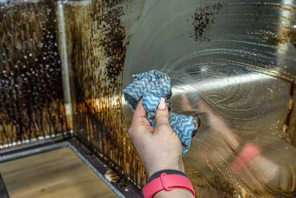 Eine Frau wäscht mit einem Küchentuch die rußverschmierte Eckscheibe eines modernen Kamins mit geschlossener Brennkammer. - Foto, Bild