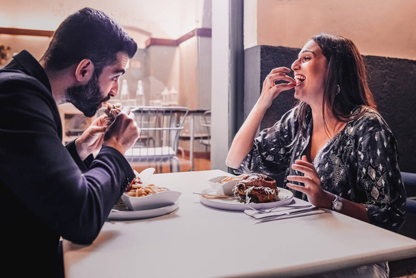 Couple of best friends having fun at the restaurant joking and eating sandwiches in the night. Engaged people dining at outdoors restaurant. Lifestyle concept. - 写真・画像