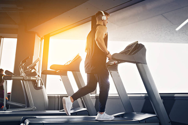Concentrated fit man listening to music in headphones while running on treadmill in modern gym with panoramic windows. - Φωτογραφία, εικόνα
