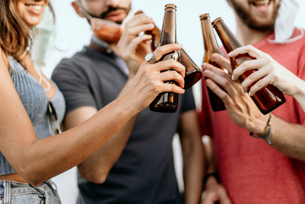 New normal lifestyle concept of young people hangout toasting with beer bottles outdoors during coronavirus breakdown wearing lowered face masks - Photo, Image