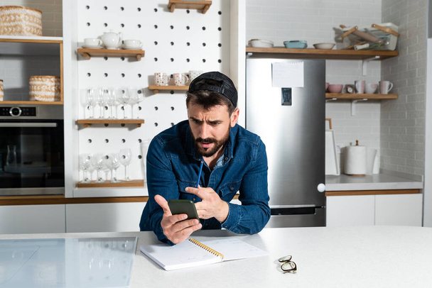 Young freelance business man in casual clothing standing at his kitchen at his home and checking the plans and statistic on his cell phone application, work at home concept - Valokuva, kuva
