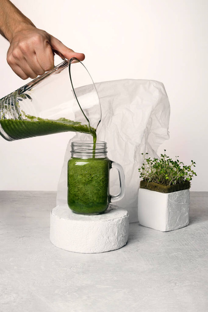 Microgreen cocktail on concrete podiums. A man's hand pours a microgreen cocktail into a glass. On a white and concrete background. - Photo, Image