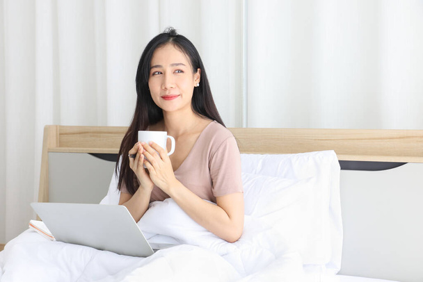 彼女のベッドの上でコーヒーを飲みながら、ラップトップで自宅からオンラインで働くアジアの女性は、 covid-19から隔離期間中に新しい通常の生活のために - 写真・画像