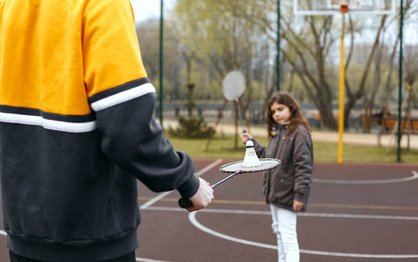 Mała kręcona dziewczynka z długimi włosami grająca w badmintona ze starszym bratem - Zdjęcie, obraz