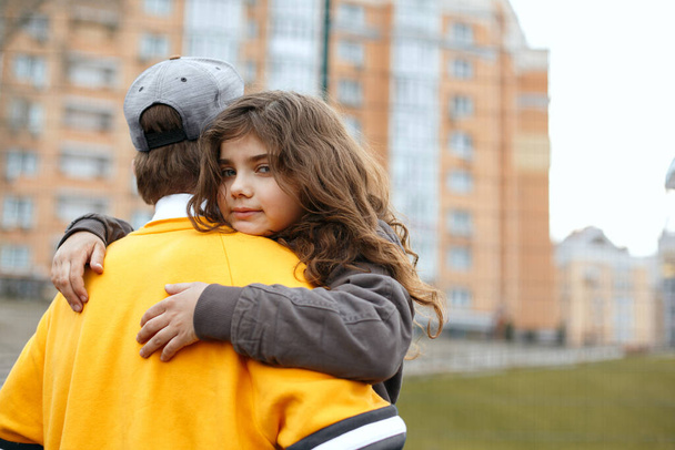 Υπέροχο σγουρό μελαχρινό κορίτσι αγκαλιάζει το μεγαλύτερο αδελφό της κατά τη διάρκεια του παιχνιδιού στην αυλή του αθλητισμού - Φωτογραφία, εικόνα