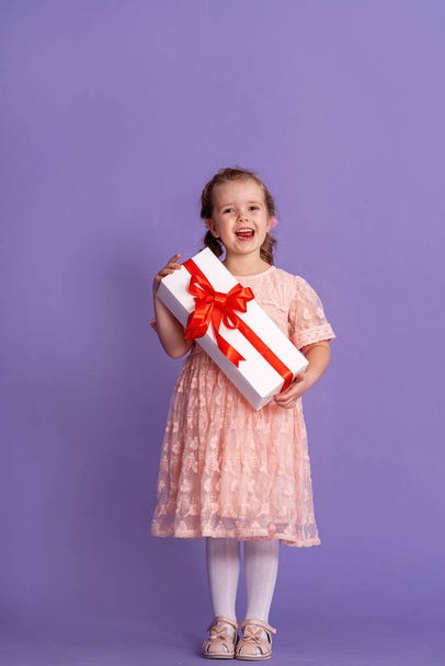 紫の背景に赤いリボンのギフトボックスのかわいい女の子。小さな子供の誕生日。幸せな子供は自分の手に贈り物を持っている。誕生日プレゼントだ。祝う、与える、受け取るという概念は - 写真・画像
