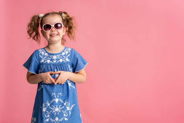 portrait of a happy cute little girl making a heart gesture, isolated on a pink studio background. Smiling baby a child with wavy hair 5 years old a charming contented cutie shows a sign of love. - Zdjęcie, obraz