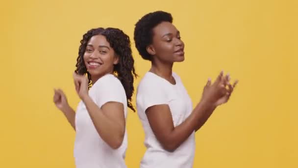 Δύο χαρούμενες αφροαμερικανίδες φίλες χορεύουν και τραγουδούν μαζί, στέκονται πλάτη με πλάτη, πορτοκαλί φόντο - Πλάνα, βίντεο