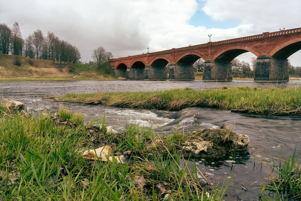 Vista de primavera del río Venta y el viejo puente de ladrillo Kuldiga con un cielo gris y nublado. - Foto, imagen