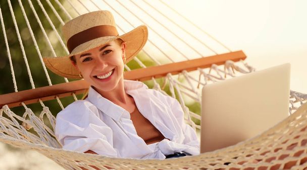 Disfrutando del trabajo durante las vacaciones de verano. Joven mujer feliz, mujer freelancer exitosa en sombrero de paja usando el ordenador portátil y sonriendo a la cámara mientras se relaja en la hamaca en la playa tropical. Trabajo a distancia - Foto, imagen