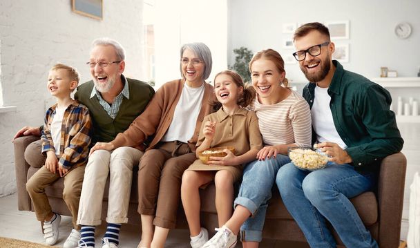 Счастливая большая семья, проводящая выходные вместе дома. Бабушка и дедушка, мама, отец и двое маленьких детей, сестра и брат, сидят на диване и едят попкорн во время просмотра смешного кино комедии по телевизору - Фото, изображение