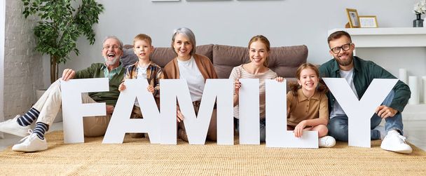 Housing and mortgage concept. Big happy family of six people grandparents, mother and father with little kids holding word FAMILY smiling at camera while sitting on floor  near sofa in the living room in new home - Фото, изображение