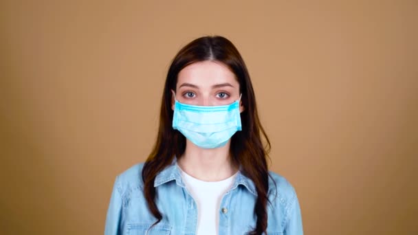 Young woman in a medical mask. The female looks seriously into the camera, shows her feet with her hands, shakes her head negatively. Health care and medical concept. Close up portrait. 4k - Footage, Video