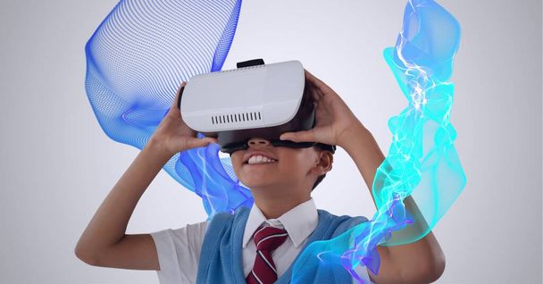 Composition of smiling schoolboy wearing vr headset with blue light trails on white background. global connection, virtual reality and technology concept digitally generated image. - Фото, зображення