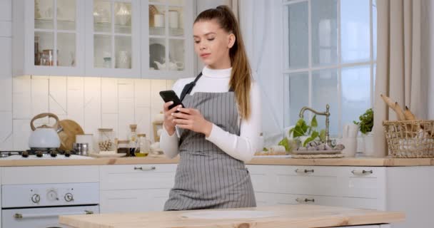 Ricette online. Giovane casalinga caucasica indossa grembiule in piedi in cucina e rete su smartphone - Filmati, video