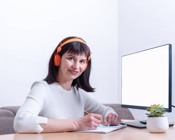 Mulher de negócios sorridente em fones de ouvido olha para você enquanto está sentado na mesa na frente de um monitor com uma tela em branco - Foto, Imagem