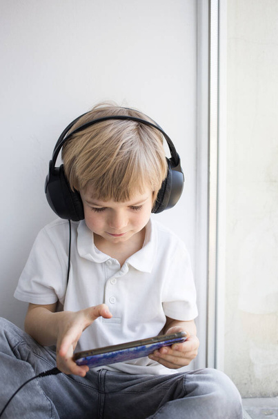 cute toddler boy in big headphones and a mobile phone, sits alone, plays games, watches cartoons or listens to music on a smartphone. Digital generation and telephone addiction concept - Foto, imagen