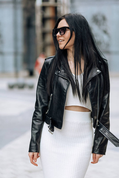 A young beautiful brunette in sunglasses and a fashionable outfit is walking around the city. Smiling portrait - Foto, imagen