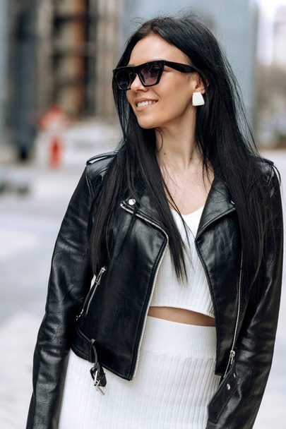 A young beautiful brunette in sunglasses and a fashionable outfit is walking around the city. Smiling portrait - 写真・画像