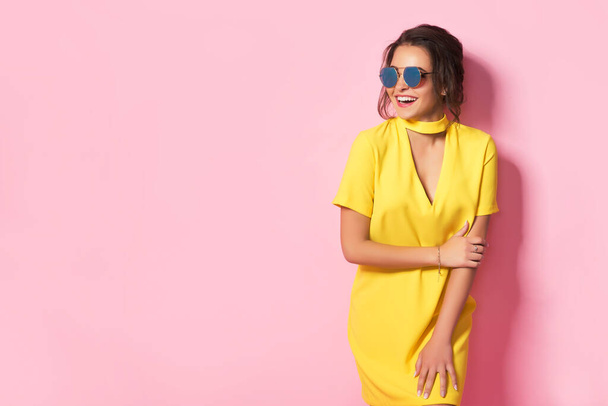 Beautiful girl in yellow dress wearing sunglasses posing, smiling on pink background in studio.  - Foto, Imagen