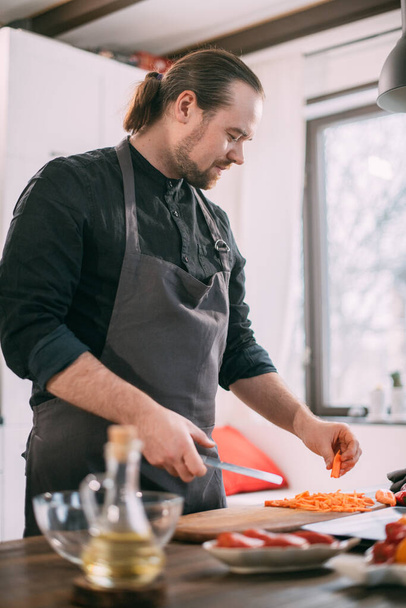 Ένας αρσενικός μάγειρας ετοιμάζει λαχανικά στο σπίτι στην κουζίνα. Νεαρός με ποδιά στην κουζίνα του σπιτιού στο τραπέζι με υλικά για βραστό λαχανικών ή σαλάτα - Φωτογραφία, εικόνα