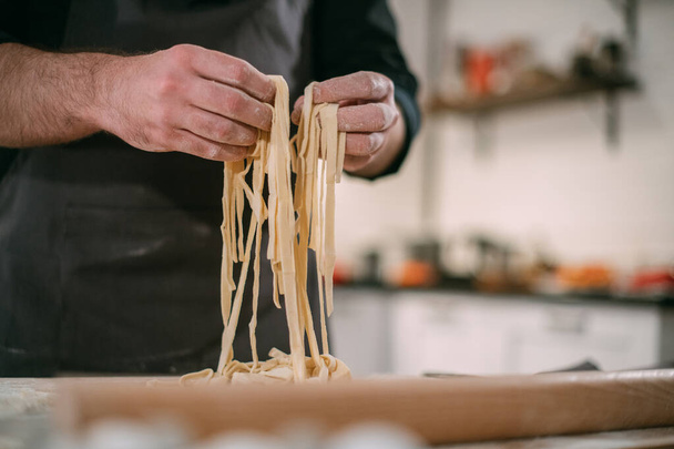 A male chef prepares noodles at home in the kitchen. Hands with rolling pin and dough. Young guy in an apron in the kitchen at the house at the table with ingredients for homemade noodles - Fotoğraf, Görsel