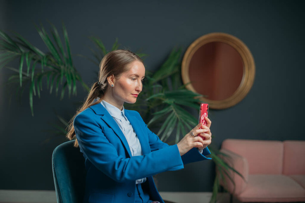 Επιχειρηματίας πορτρέτο γυναίκα στο σύγχρονο εσωτερικό του γραφείου. Χαμογελώντας κομψό νεαρό κορίτσι σε ένα μπλε κοστούμι επιχείρησης κάνει μια selfie στο τηλέφωνο, νοικοκυρεύεται. Γυναίκα στις επιχειρήσεις, πορτρέτο μιας επιχειρηματία. - Φωτογραφία, εικόνα