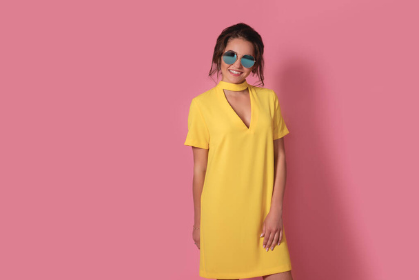Beautiful girl in yellow dress wearing sunglasses posing, smiling on pink background in studio.  - Фото, изображение
