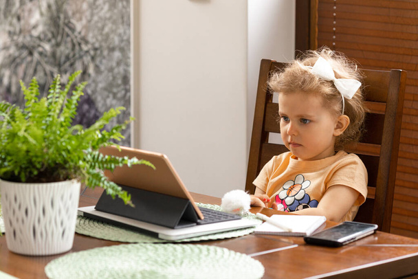 Vauva tyttö 3-vuotias kihara tukka istuu pöydässä. Online Kodinhoito tabletilla kotona. Koulutuslapsen käsite. Coronavirus. - Valokuva, kuva