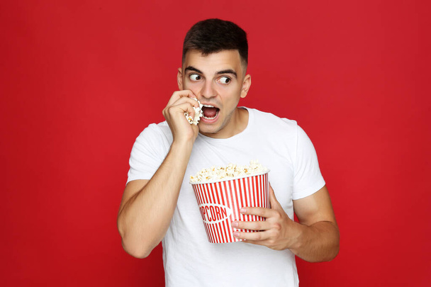 Young man with bucket of popcorn on red background - Фото, изображение