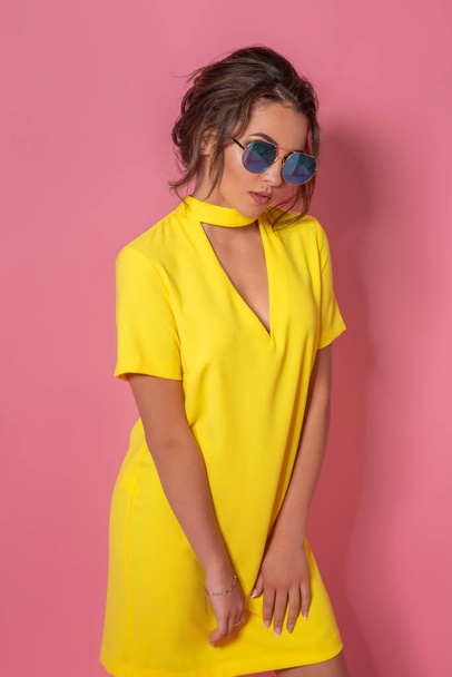 Beautiful girl in yellow dress wearing sunglasses posing, smiling on pink background in studio.  - Foto, afbeelding