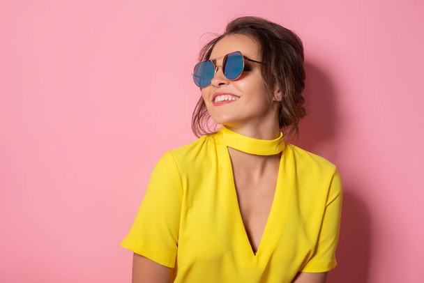 Belle fille en robe jaune portant des lunettes de soleil posant, souriant sur fond rose en studio.  - Photo, image