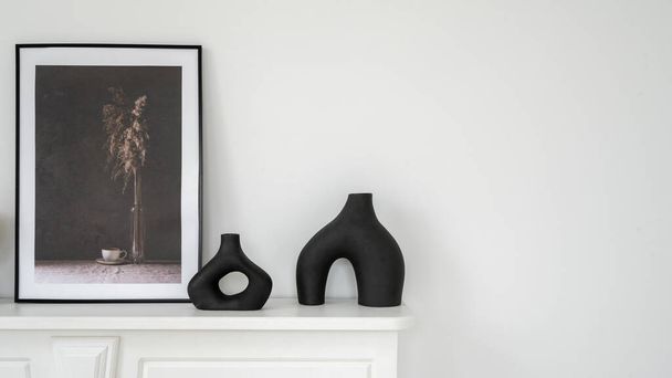 家の装飾の概念。暖炉の上の棚の上にフレーム内のエレガントな黒い花瓶やモックアップ画像のパノラマビュー。現代的なインテリアデザインと白のコピースペースの壁と新しいアパート - 写真・画像