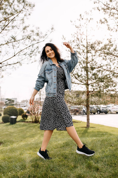 Happy young brunette woman, wearing a denim jacket and a dress, walking in a park. - Photo, Image