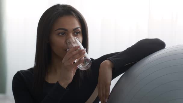 Африканская женщина пьет воду во время тренировки сидя рядом с Фитболом в помещении - Кадры, видео
