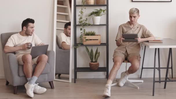 Széles felvétel fiatal szőke fehér férfi ül az íróasztalnál otthon, videó digitális tabletta segítségével vezeték nélküli fülhallgató, majd beszél a barátja dolgozik laptop a közelben - Felvétel, videó