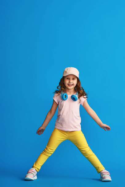 little cute girl in fashionable summer clothes and headphones stands on a blue background in full growth. The concept of summer and spring casual children's clothing. - Foto, Bild