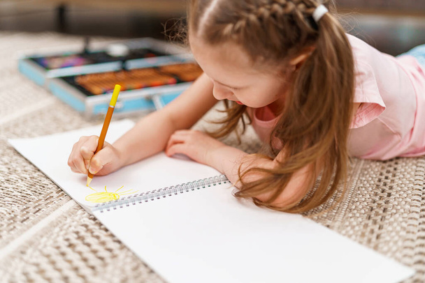 Little cute Caucasian girl child, drawing in an album with colored pencils, lying on floor. hardworking child does homework. The child's hobby is drawing. new modern set for drawing and creativity - Fotoğraf, Görsel