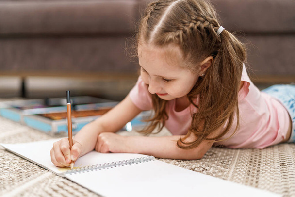Little cute Caucasian girl child, drawing in an album with colored pencils, lying on floor. hardworking child does homework. The child's hobby is drawing. new modern set for drawing and creativity - Φωτογραφία, εικόνα