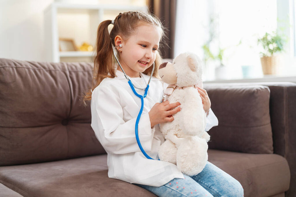 little Caucasian girl in glasses and a white medical uniform is treating a teddy bear, playing doctor and hospital. A happy child acts as a doctor examining a toy in a veterinary clinic. - Foto, Bild