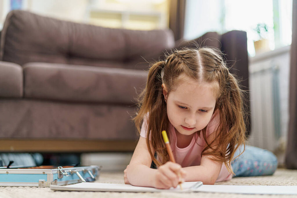 Little cute Caucasian girl child, drawing in an album with colored pencils, lying on floor. hardworking child does homework. The child's hobby is drawing. new modern set for drawing and creativity - 写真・画像