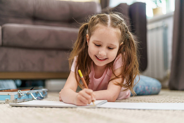 Little cute Caucasian girl child, drawing in an album with colored pencils, lying on floor. hardworking child does homework. The child's hobby is drawing. new modern set for drawing and creativity - Foto, imagen
