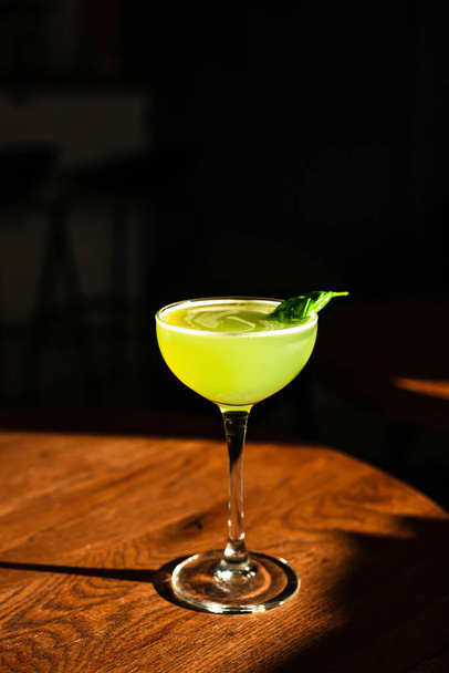 A sweet and sour refreshing basil gin sour cocktail in a coupe glass in direct hard sunlight with long shadows, on a wooden table, vertical photo with copy space - Фото, изображение