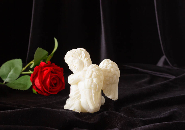 Una pequeña escultura de un angelito con alas y una rosa roja sobre un fondo negro. El concepto de funerales, condolencias y luto. - Foto, Imagen